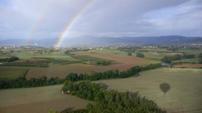 Links im Bild ein Regenbogen über der Landschaft des Zentralmassivs, rechts unten der Schatten des Heißluftballons.
